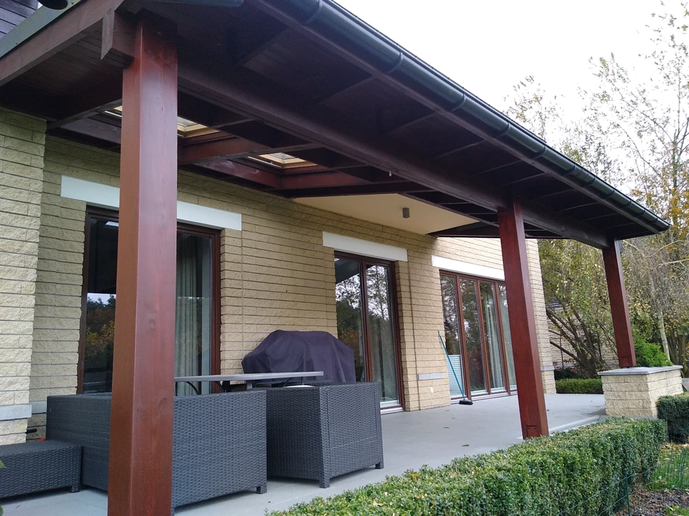 Montaż rynien na drewnianym zadaszeniu tarasu przy elewacji z tynkowanych bloczków i meblach ogrodowych